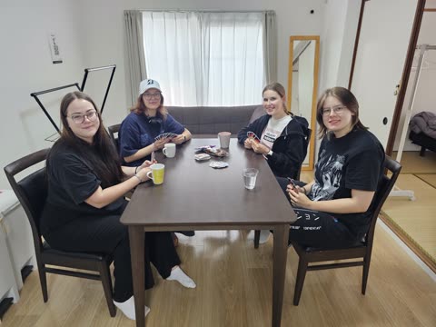 Four young women sit around a dark wooden table holding playing cards inside a living room.