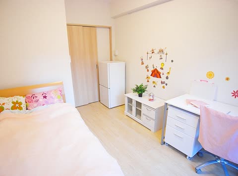 A brightly lit bedroom with a bed, white desk, small refrigerator, and circus-themed wall decals on a light wood floor.