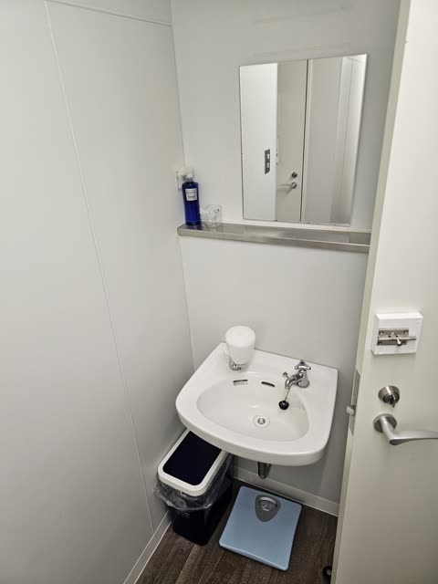 A compact washroom contains a small white sink, a mirror, and a trash can and weight scale on the dark floor.