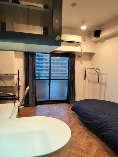 A small apartment room with herringbone wood floors, a bed with a dark blue cover, and a sliding glass door with blue curtains.