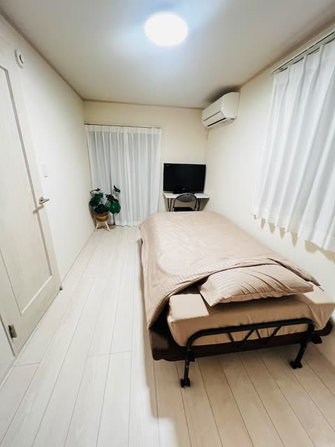 A narrow bedroom with light wood floors contains a single bed on a wheeled frame, a small desk with a TV, and a potted plant.