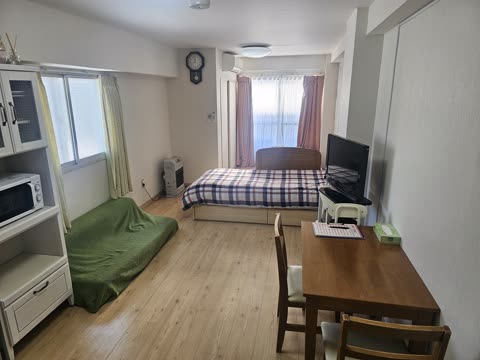 A furnished studio apartment with a bed, a green floor mattress, a wooden dining table, and a white kitchen hutch.