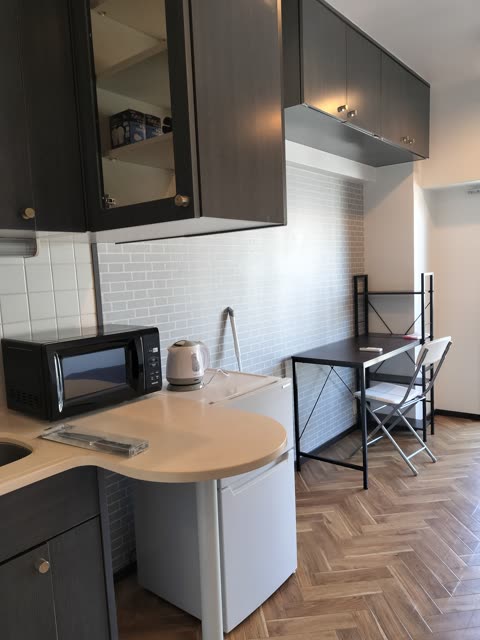 A kitchenette with dark cabinets and appliances sits next to a small desk and chair on a herringbone wood floor.