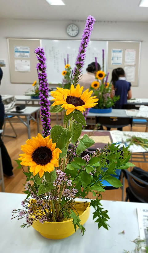 Sunflowers and purple flowers arranged in a yellow bowl on a classroom table.