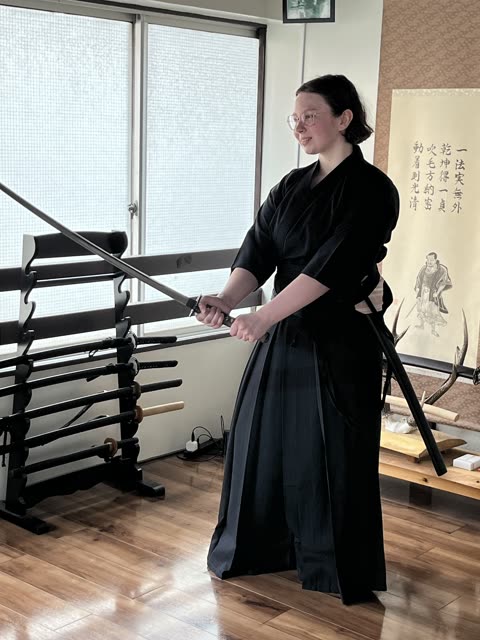 Person in black kimono handling a sword near a sword rack.