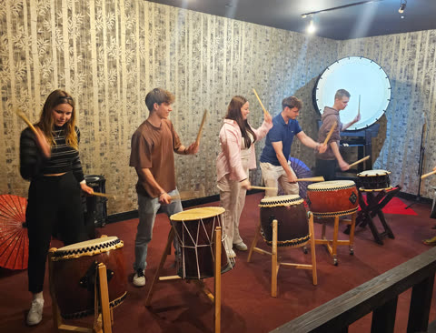 Five people stand in a row playing large taiko drums in a room with bamboo-patterned wallpaper.