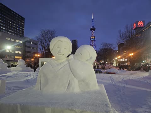 Two snow sculptures embracing at night in a snowy city square.