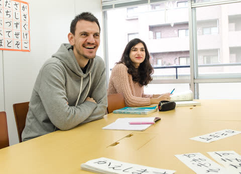 A smiling man and a woman sit at a table with books and Japanese character cards in a classroom.