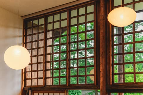 Two round paper lanterns hang in a room with large wooden grid windows that look out onto green trees.