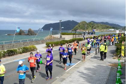 Runners race along a coastal road.