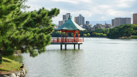 Red gazebo sits on a lake, city buildings in the background.