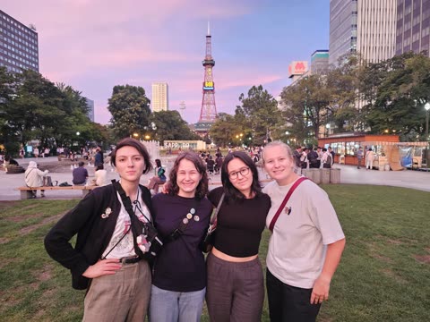 Four women stand together smiling in a city park at dusk with the Sapporo TV Tower in the background.