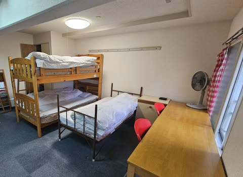 A dorm room contains a wooden bunk bed, a single bed, and a wooden desk with red chairs on blue carpeting.