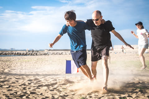 Two men run on sandy beach, one supporting the other near a French flag.
