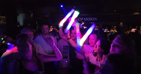 People holding illuminated glow sticks dance in a dark nightclub.