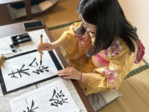 A person in a yellow floral yukata kneels at a low table, painting Japanese characters with a brush.