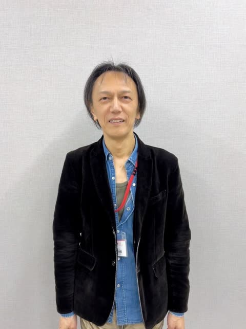 A man with shoulder-length dark hair in a black jacket and denim shirt smiles while standing in front of a gray wall.