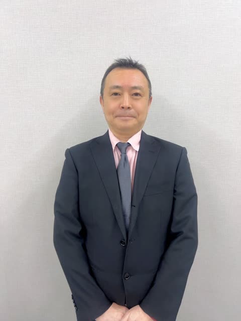 A man in a black suit, pink shirt, and grey tie stands with his hands clasped in front of a textured grey wall.
