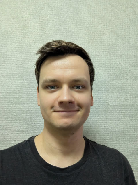 A man with short brown hair and blue eyes looks at the camera with a slight smile in front of a plain, light-colored wall.