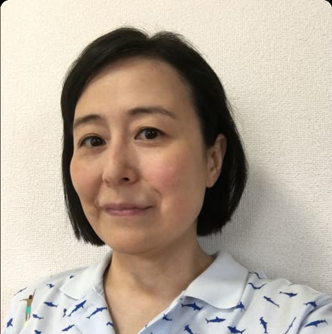 A woman with short dark hair wearing a light blue polo with a shark pattern smiles in front of a textured white wall.