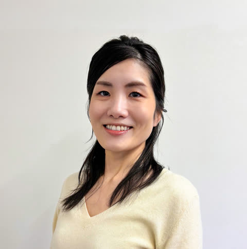 A woman with dark hair and a cream-colored sweater smiles while looking forward against a plain white wall.