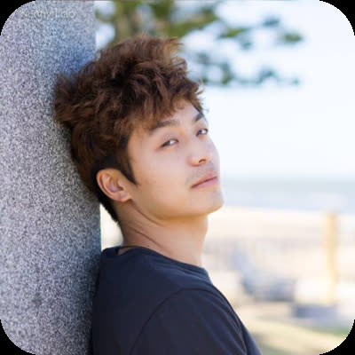 A man with styled brown hair leans against a grey stone pillar and looks over his shoulder outdoors.