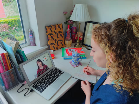 Woman writes in notebook at desk, while watching laptop screen.