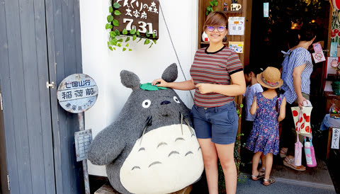 A woman in a striped shirt stands smiling with her hand on a large, grey, furry character outside a shop entrance.